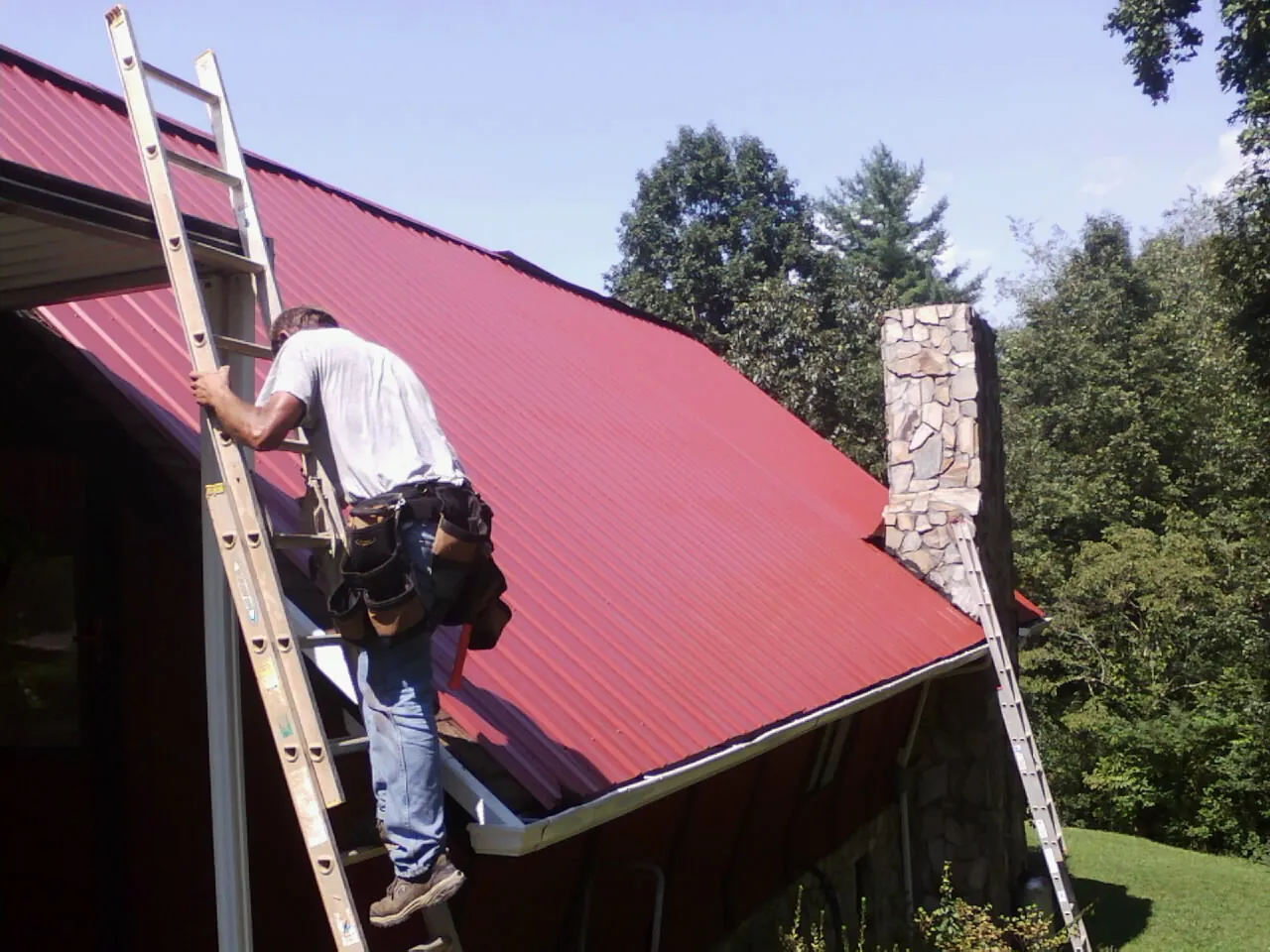 Roofer on a ladder beside a red metal roof and stone chimney — Soffit & Fascia Repair by Roofing Brothers in Wingdale